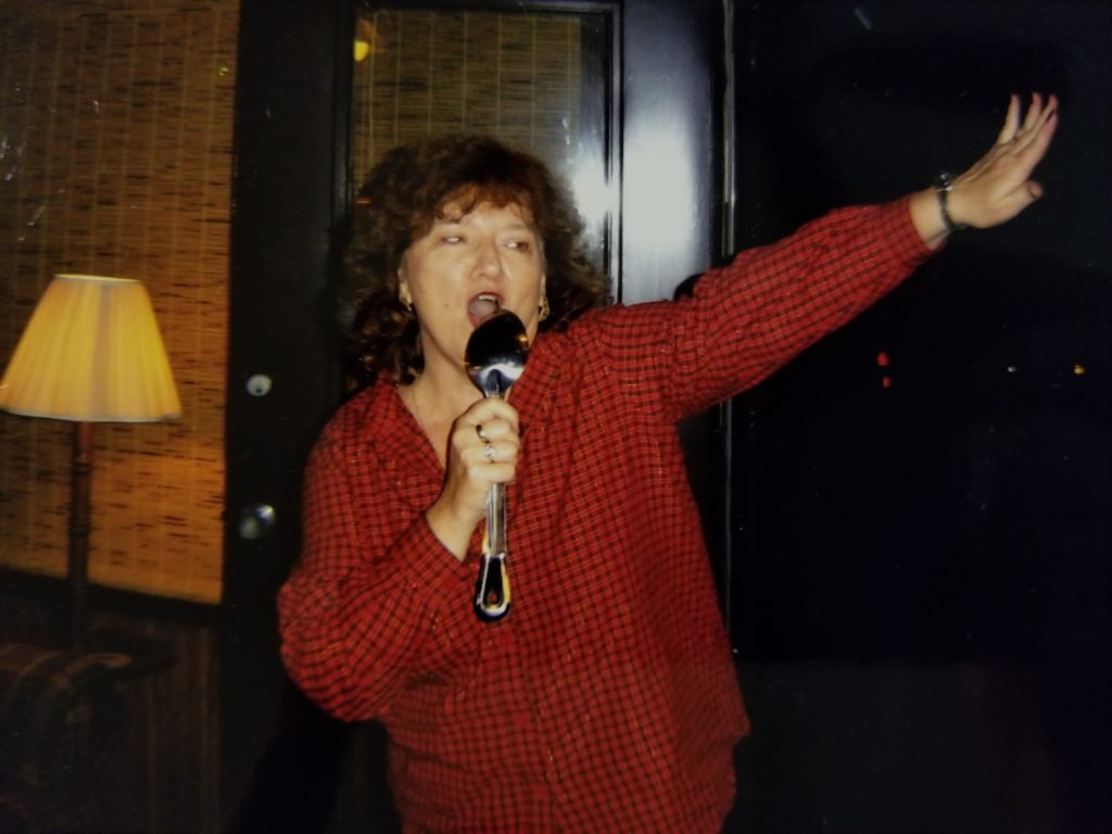 Woman in red blouse singing, holding a large soup spoon.